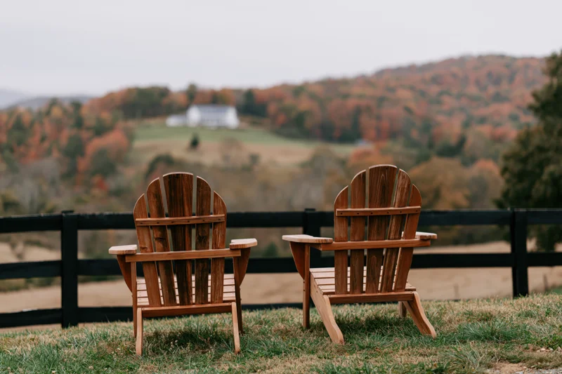 Two empty wooden adirondack chairs side by side on a grassy hill overlooking autumn hills and a distant white house