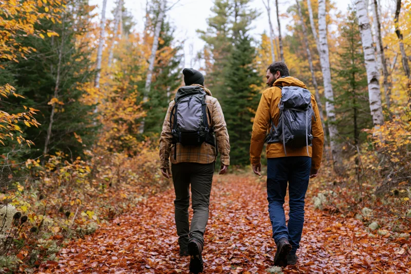 Two men with backpacks walking along a leaf-covered trail in an autumn forest