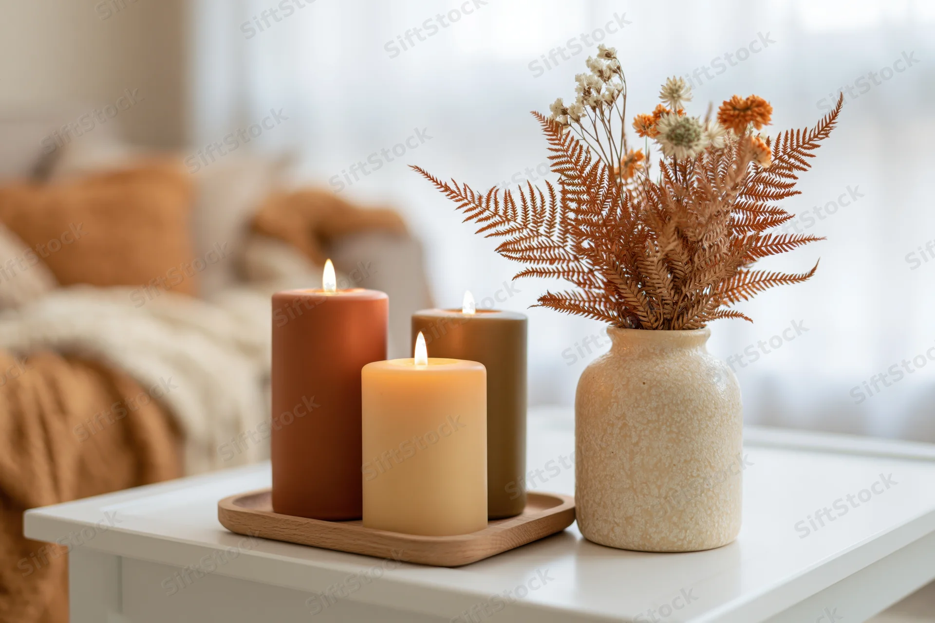 Three lit candles in terracotta and olive tones on a wooden tray beside a ceramic vase with dried ferns and flowers on a white table in a cozy living room