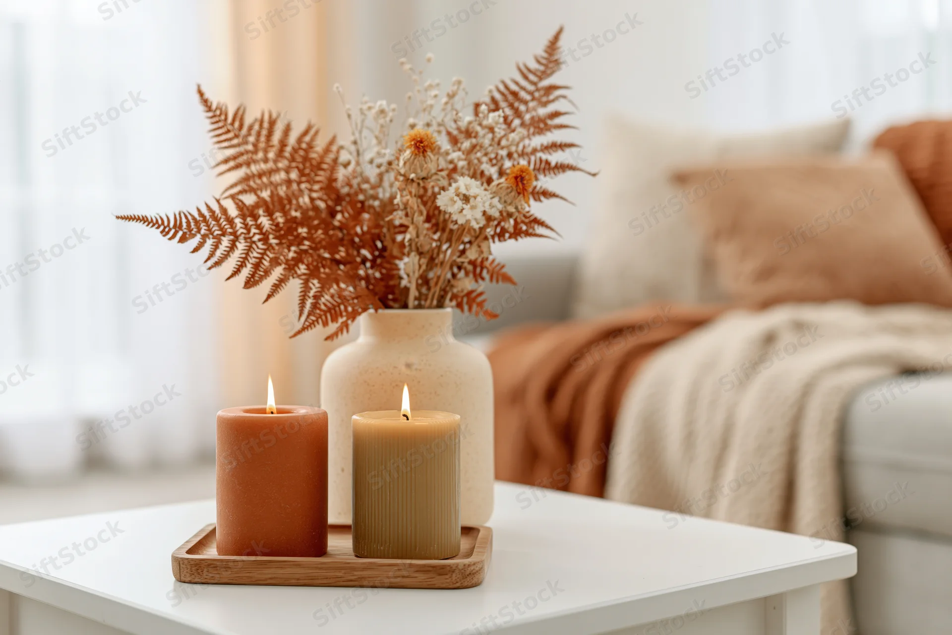 Two lit candles in terracotta and olive tones on a wooden tray with a ceramic vase of dried ferns and flowers on a white coffee table in a cozy living room