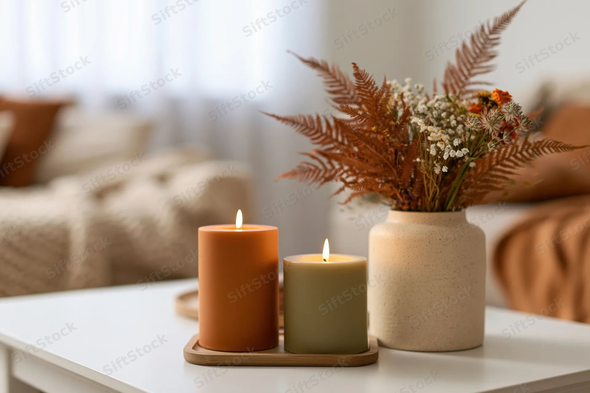 Two lit candles in terracotta and olive tones on a tray beside a vase of dried ferns and flowers on a white table in a living room