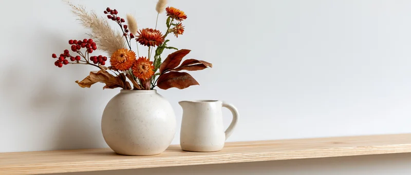 Dried flowers and berries in a white ceramic vase beside a pitcher on a light wood shelf