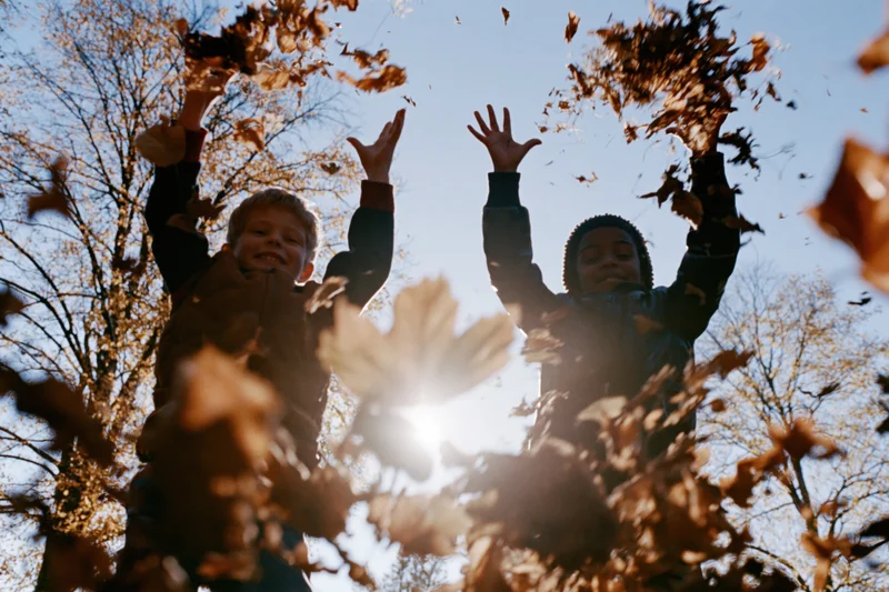 Two children jumping and tossing autumn leaves in a sunlit park