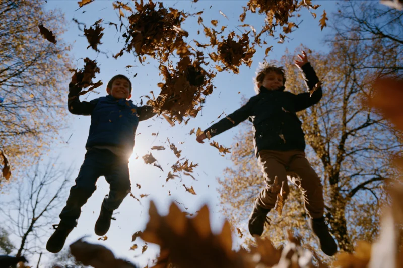 Two boys jumping in midair tossing autumn leaves in a park under sunlight