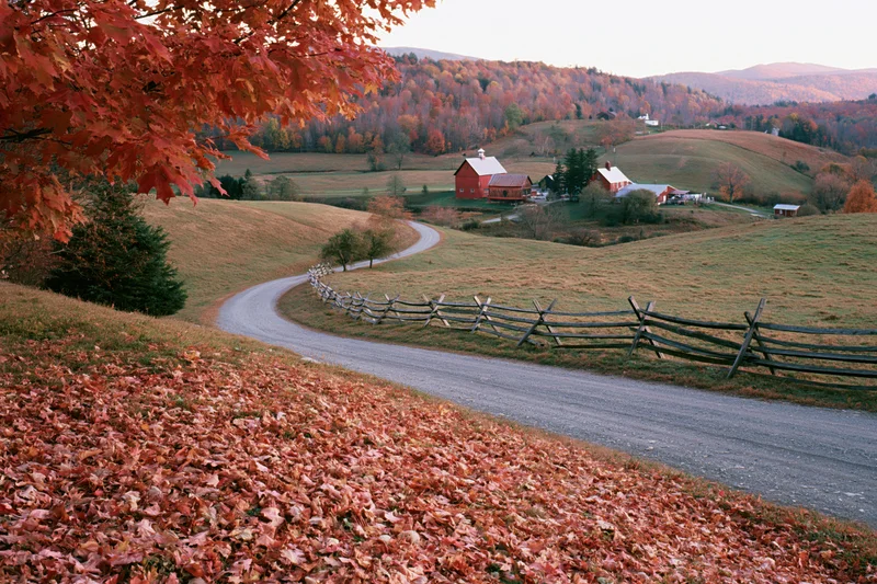 Winding gravel road through autumn foliage-covered hills leading to red barn farm in Vermont countryside