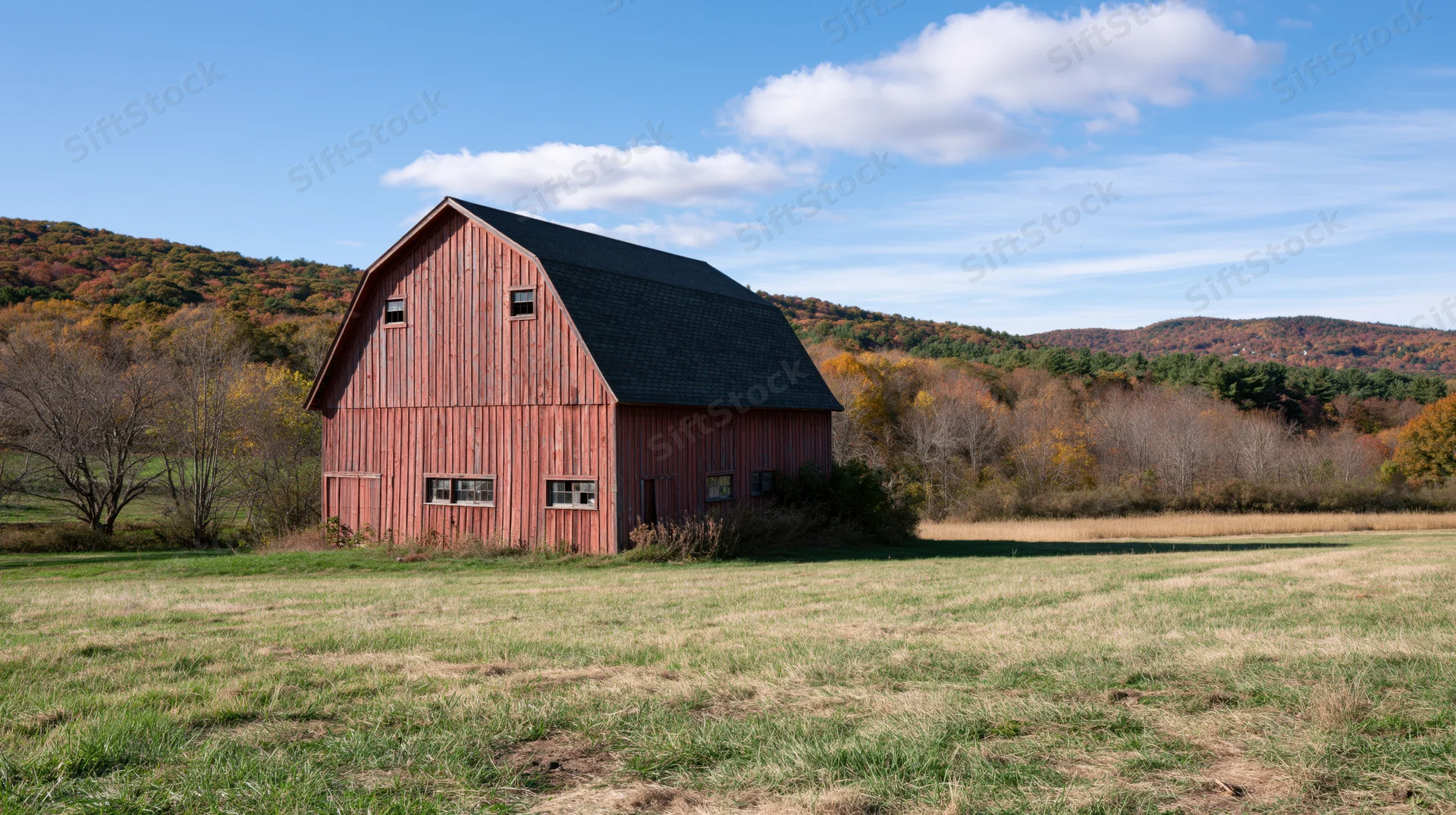 Red barn with dark gambrel roof standing in open field surrounded by autumn trees under blue sky with clouds