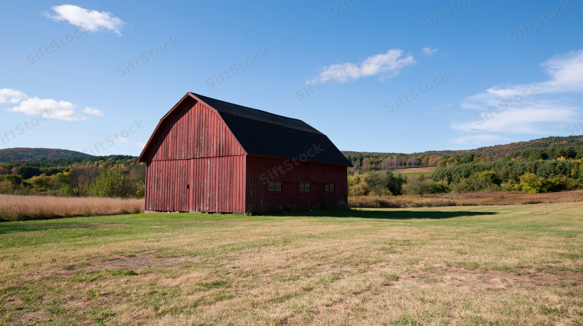 Weathered red barn with dark gambrel roof standing in open field under blue sky