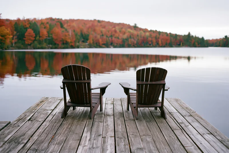 Two adirondack chairs on a weathered wooden dock facing a calm lake with autumn foliage reflected in the water