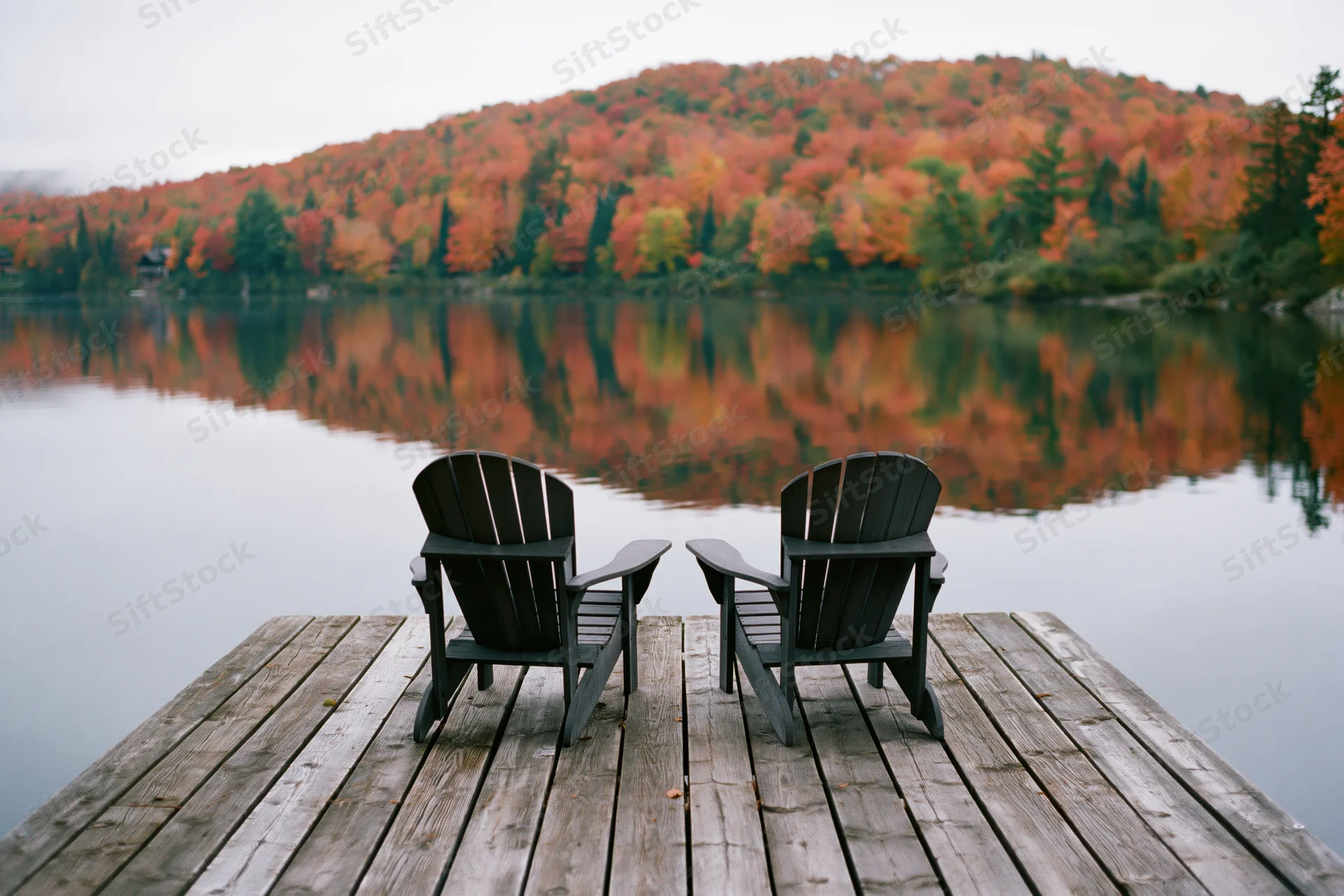 Two adirondack chairs on a weathered wooden dock facing a calm lake with autumn forest reflection