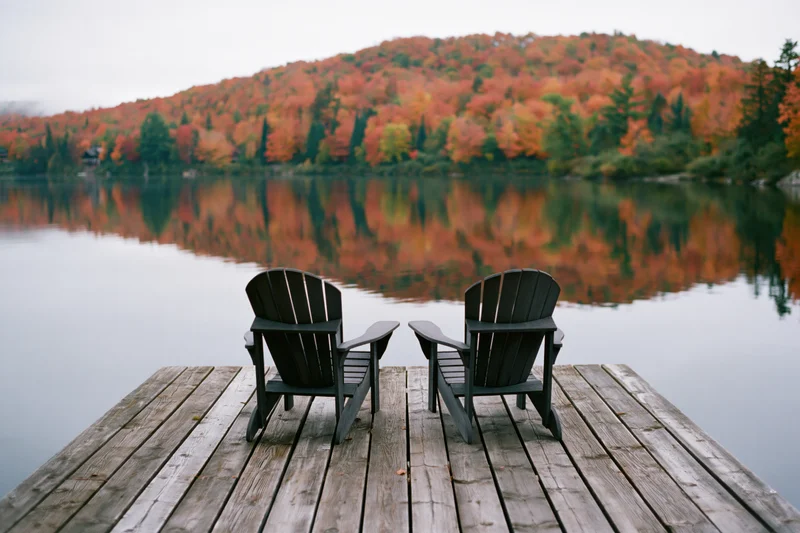 Two adirondack chairs on a weathered wooden dock facing a calm lake with autumn forest reflection