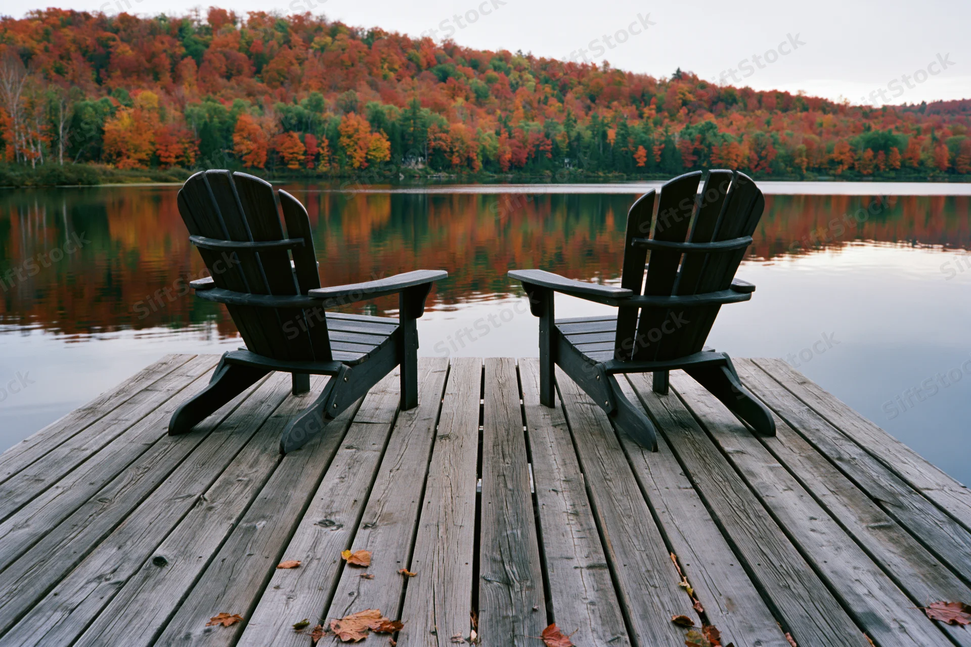Two Adirondack chairs on a weathered wooden dock facing a calm lake with autumn forest reflection