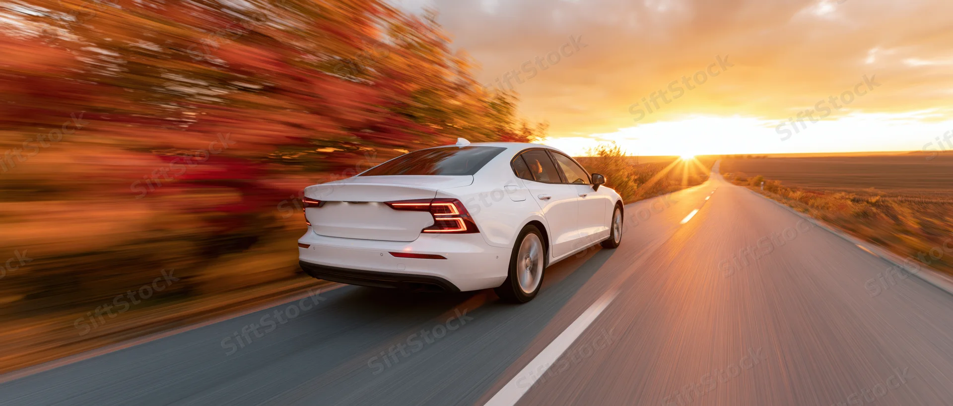 White sedan driving away on a straight country road at sunset