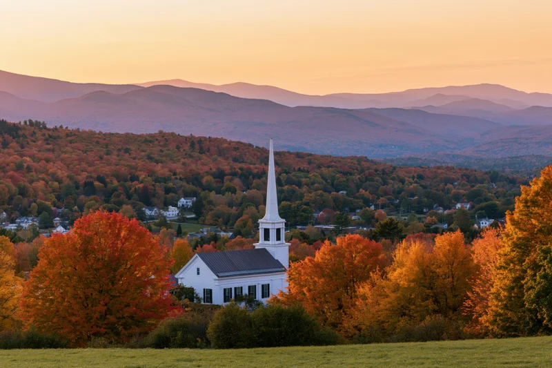 White church with steeple rising above autumn trees in a rural valley at sunset
