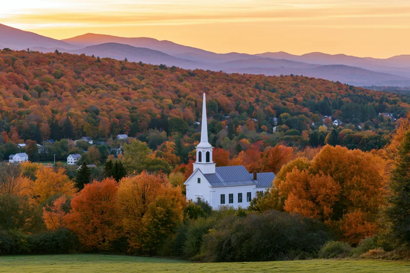 White new england church with steeple surrounded by autumn foliage in a rural valley at sunset