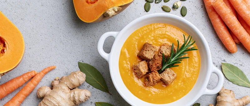 Creamy butternut squash soup in a white ceramic bowl with croutons and rosemary, surrounded by carrots, ginger, and squash halves on a gray surface