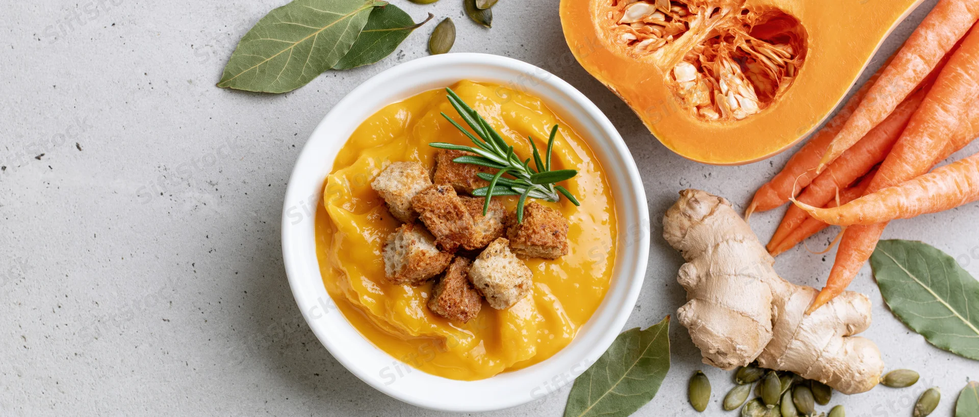 Creamy butternut squash soup in a white ceramic bowl topped with croutons and rosemary, surrounded by carrots, ginger, pumpkin seeds, and bay leaves on a gray countertop