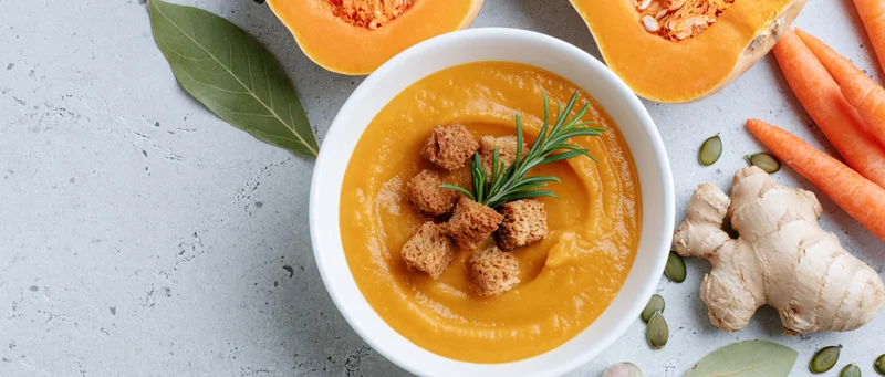 Creamy butternut squash soup in a white ceramic bowl garnished with rosemary and croutons surrounded by carrots, ginger, pumpkin seeds, and bay leaves on a gray surface