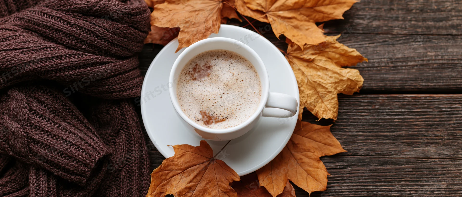 White ceramic coffee cup with froth on saucer surrounded by autumn maple leaves and brown knit sweater on wooden table