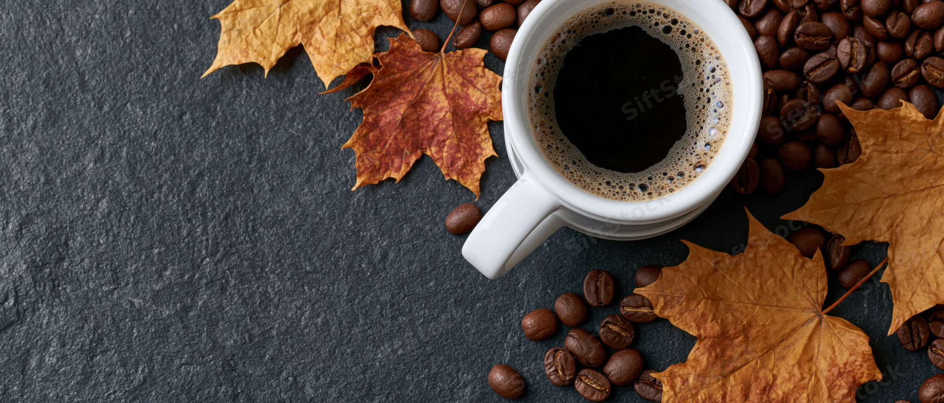 White ceramic mug filled with frothy black coffee surrounded by autumn maple leaves and coffee beans on dark slate surface