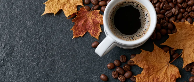 White ceramic mug filled with frothy black coffee surrounded by autumn maple leaves and coffee beans on dark slate surface