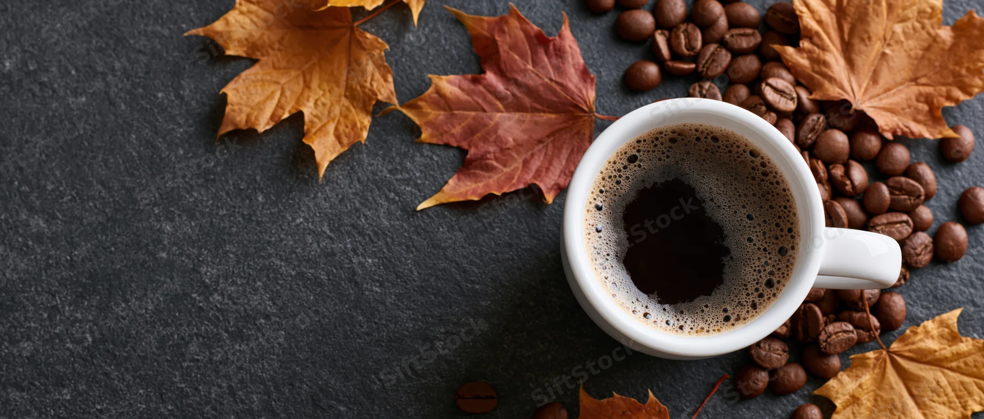 White ceramic mug filled with frothy black coffee surrounded by autumn leaves and coffee beans on dark slate surface
