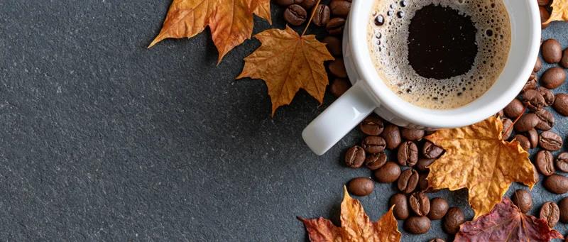 White ceramic mug filled with frothy black coffee surrounded by coffee beans and autumn maple leaves on dark stone surface