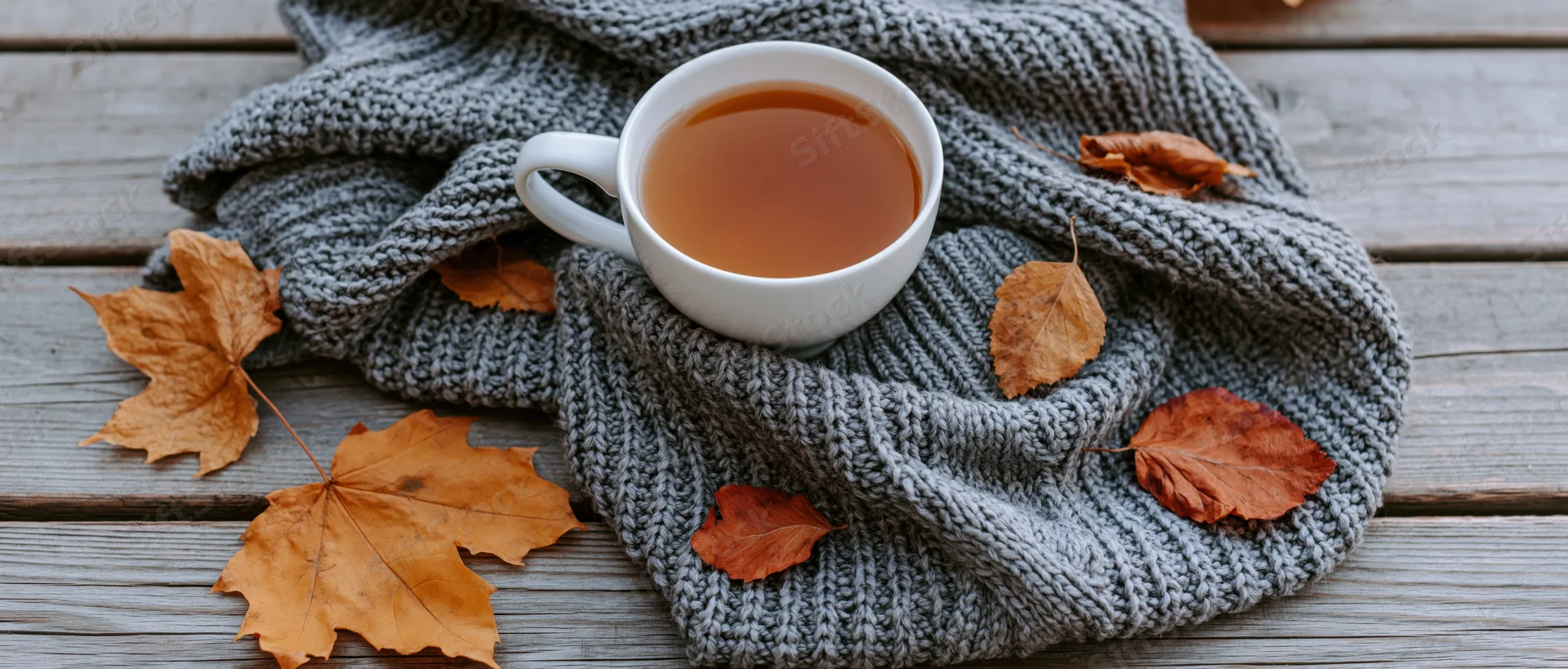 White ceramic mug of amber tea resting on a gray knitted sweater with autumn leaves on a wooden table