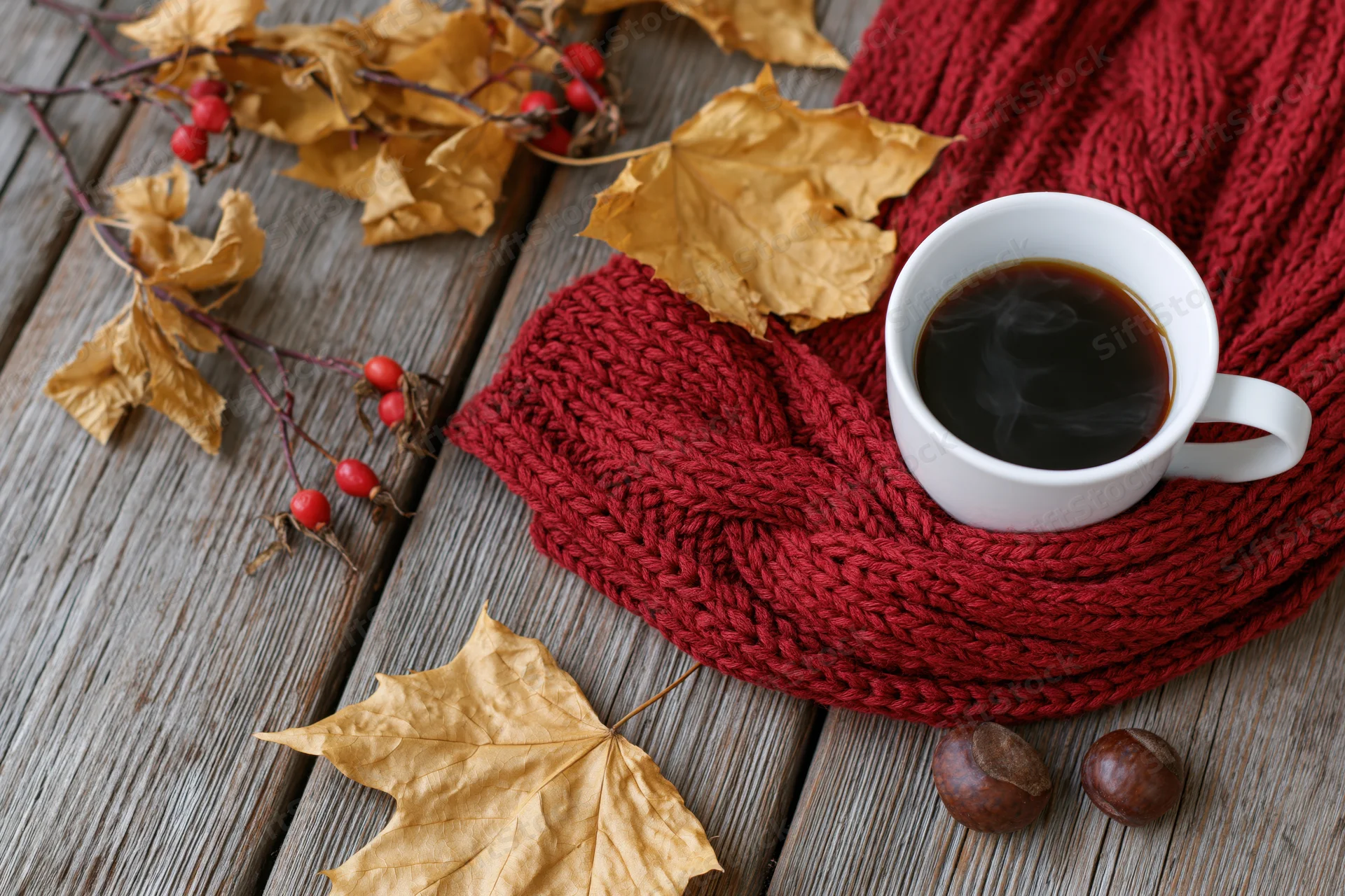 Steaming black coffee in a white ceramic mug resting on a red knitted scarf with autumn leaves and berries on a wooden table