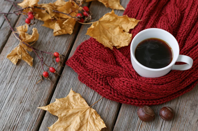 Steaming black coffee in a white ceramic mug resting on a red knitted scarf with autumn leaves and berries on a wooden table