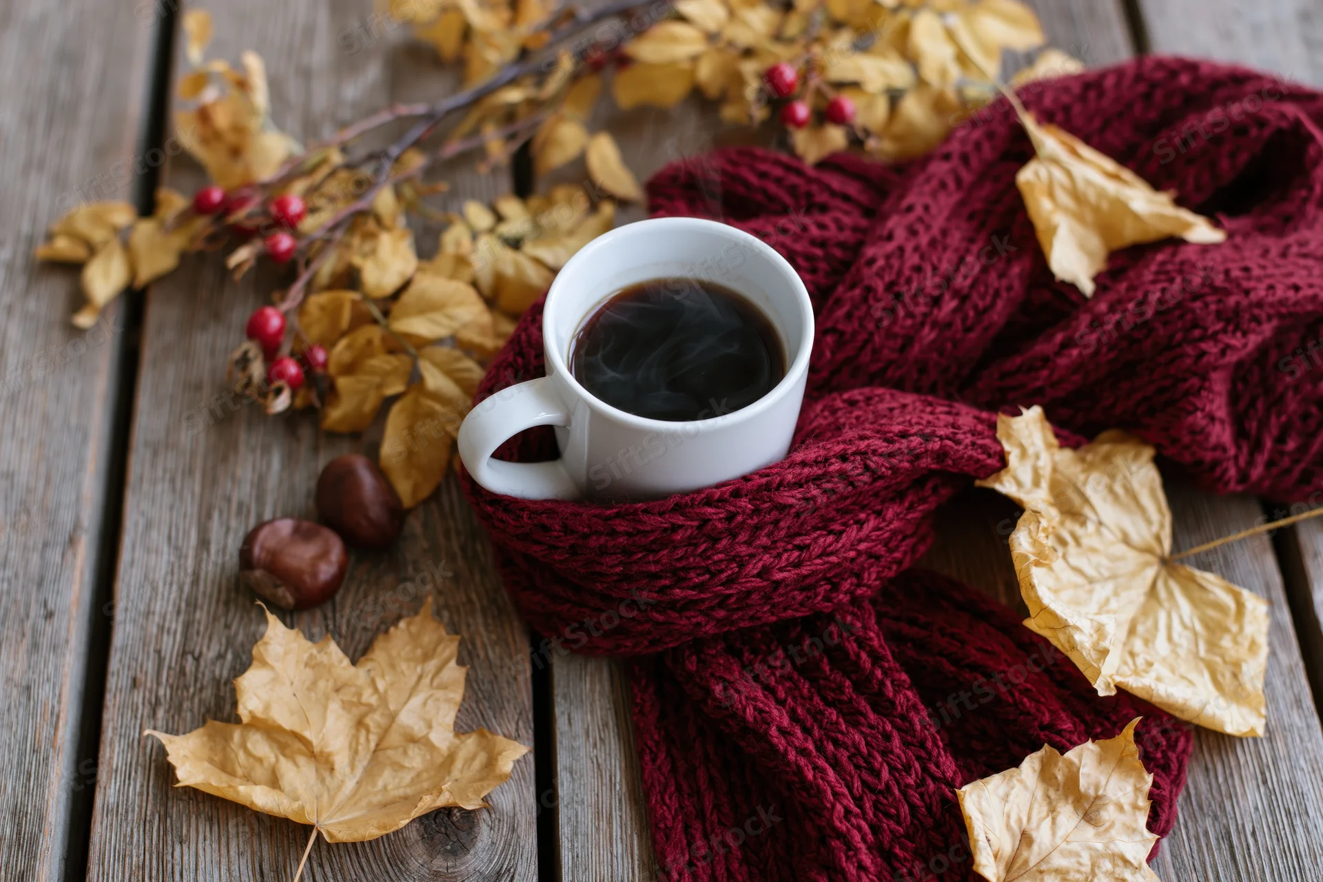Steaming coffee mug wrapped in a red knitted scarf on a wooden table with dried autumn leaves and berries