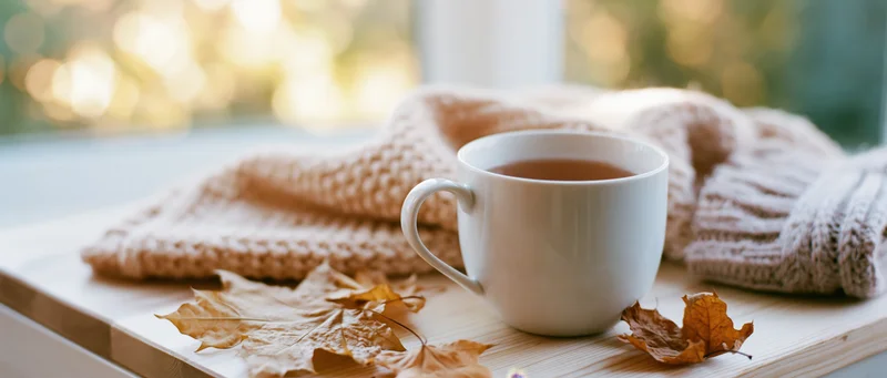White ceramic mug of tea on wooden surface beside knitted sweater and autumn leaves near window