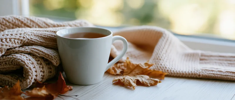 White ceramic mug of tea beside knitted sweater and autumn leaves on wooden surface near window