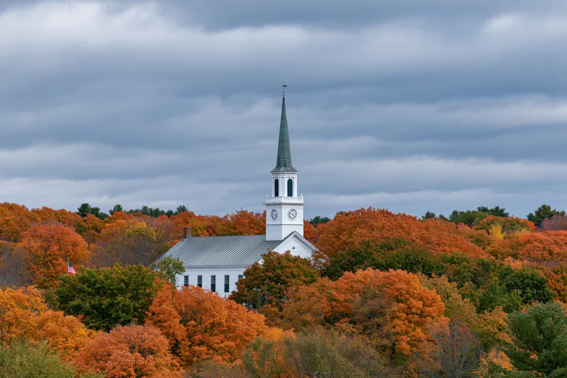 White clapboard church with green steeple and clock tower surrounded by autumn foliage under cloudy sky