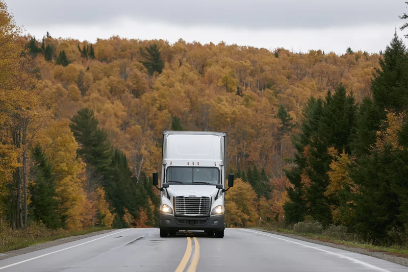 White semi-truck driving toward camera on a two-lane rural highway lined with autumn trees