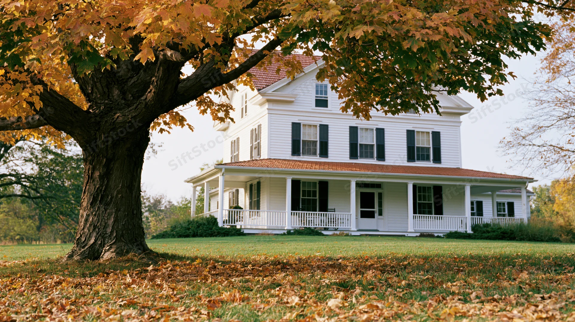 White two-story farmhouse with wraparound porch and autumn tree in front lawn