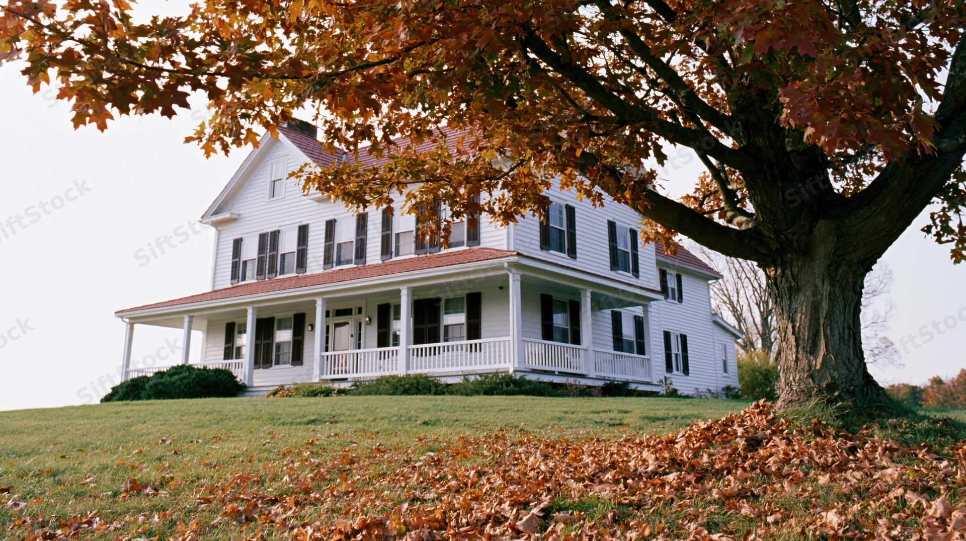 White two-story farmhouse with wraparound porch on a grassy hill in autumn