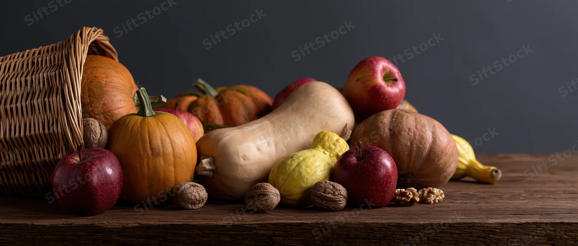 Wicker cornucopia basket tipped over spilling pumpkins, squash, apples and walnuts onto wooden table