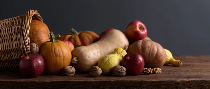 Wicker cornucopia basket tipped over spilling pumpkins, squash, apples and walnuts onto wooden table