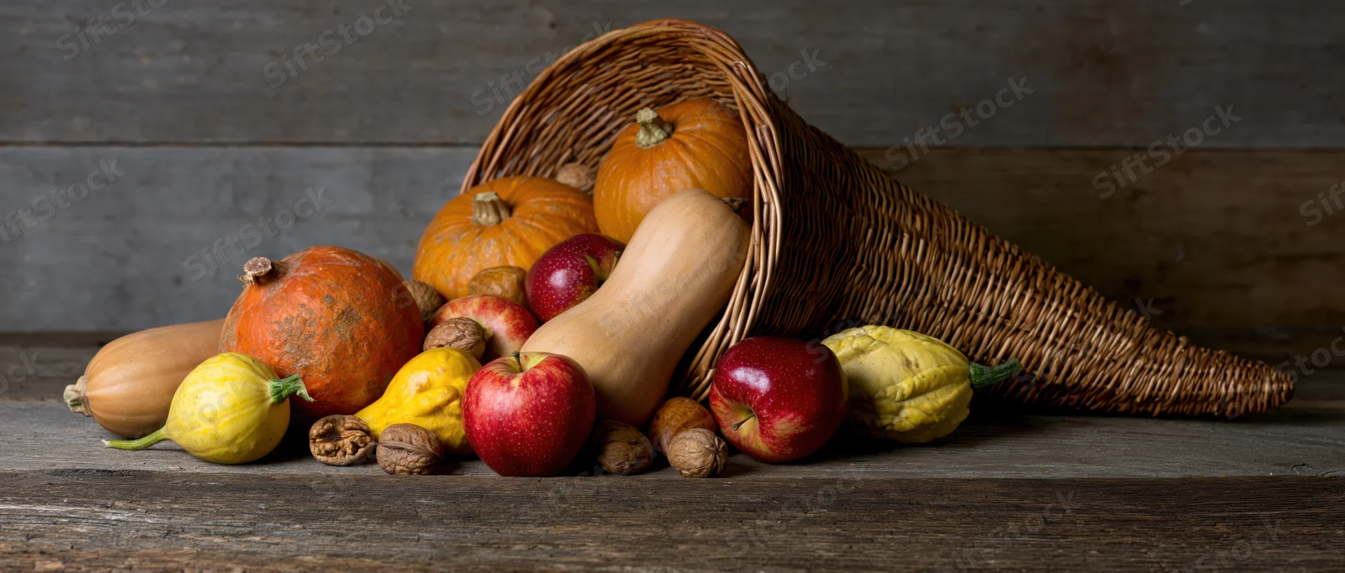 Wicker cornucopia tipped on wooden table spilling pumpkins, squash, apples and walnuts