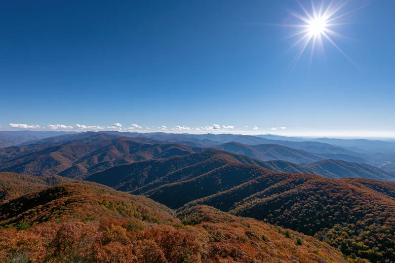 Rolling mountain ridges covered in autumn foliage under a clear blue sky with bright sun