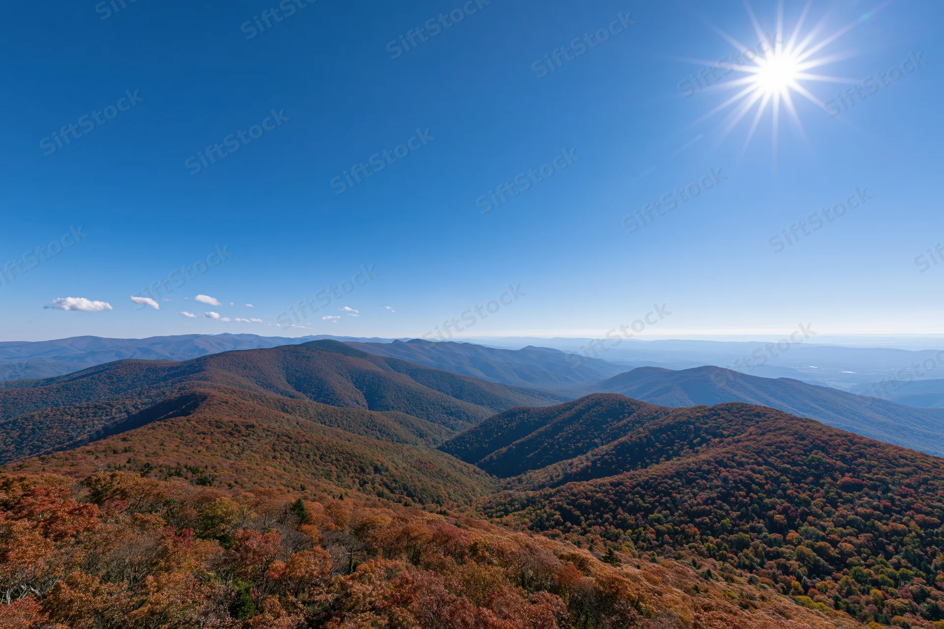 Rolling mountain ridges covered in autumn foliage under a bright blue sky with the sun shining