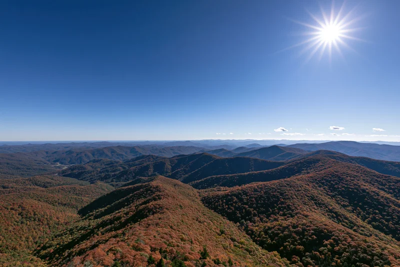 Rolling mountain ridges covered in autumn foliage under a clear blue sky with bright sun