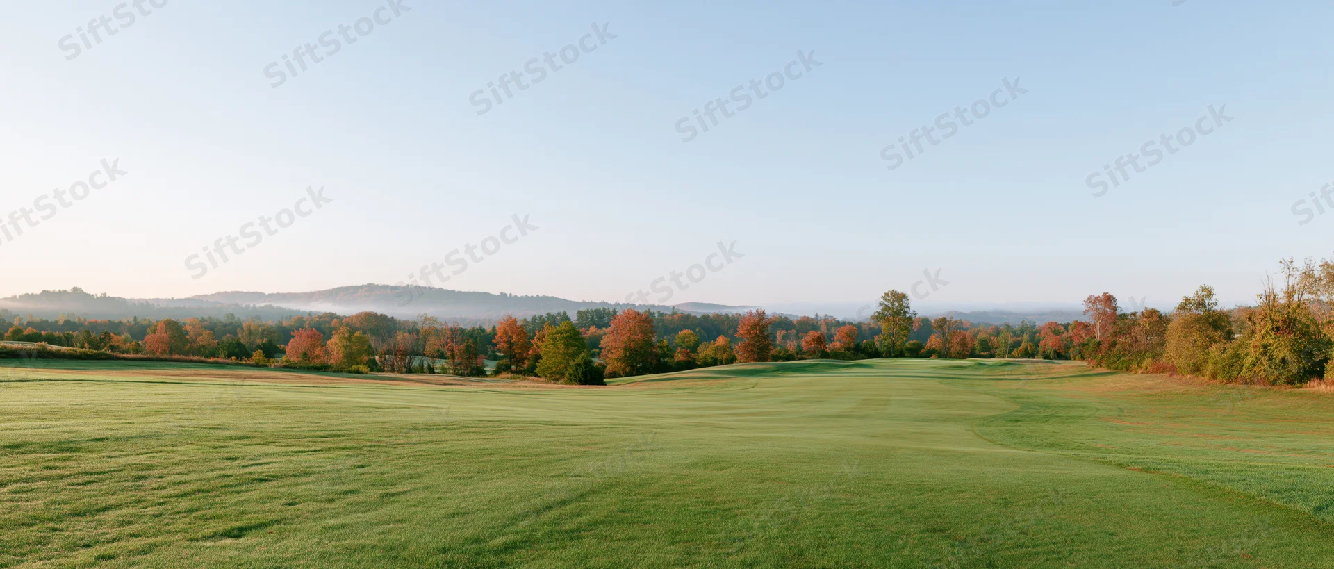 Wide panoramic view of a gently rolling golf course fairway with autumn trees and distant hills under a clear sky