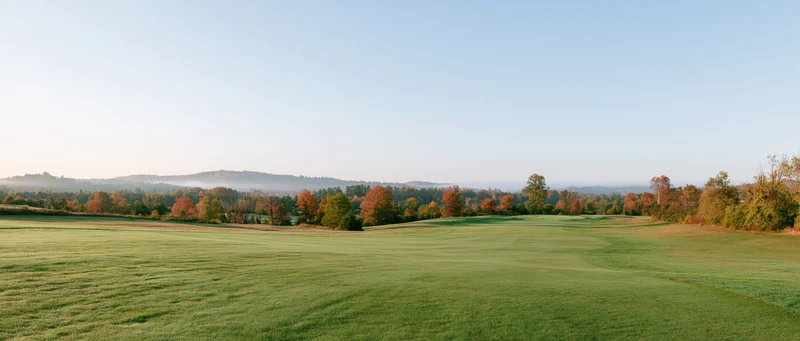Wide panoramic view of a gently rolling golf course fairway with autumn trees and distant hills under a clear sky