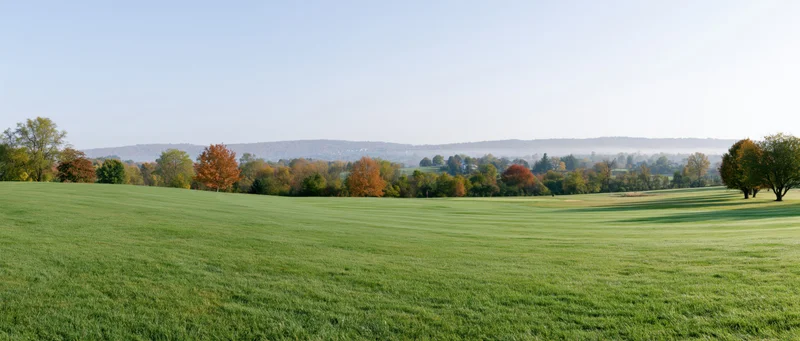 Wide panoramic view of a gently rolling golf course fairway with autumn trees and distant hills under a clear blue sky