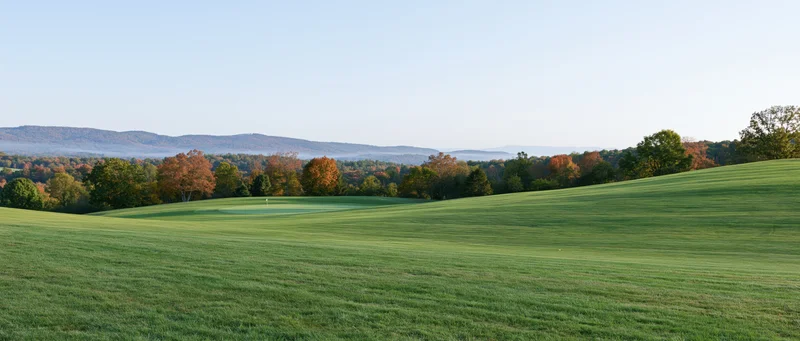 Wide panoramic view of a gently rolling golf course fairway with autumn trees and distant hills under a clear sky