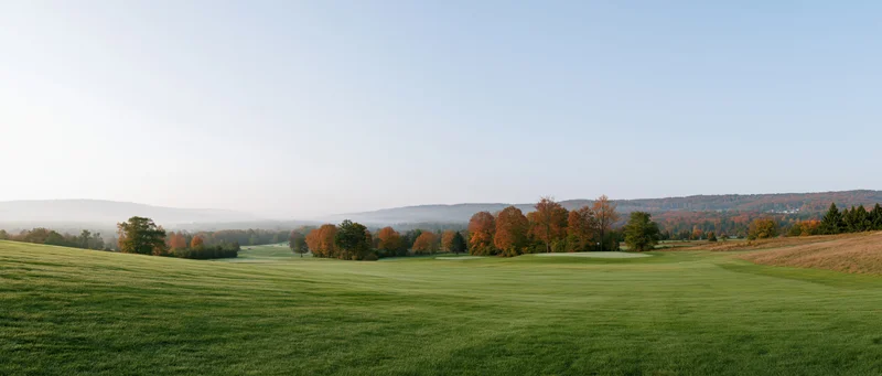 Wide panoramic view of a gently rolling golf course fairway with autumn trees and distant hills under a clear sky
