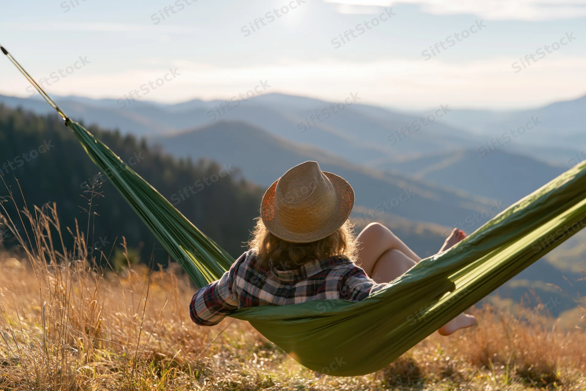 Woman reclining in green nylon hammock on mountain hill overlooking valley and forested landscape