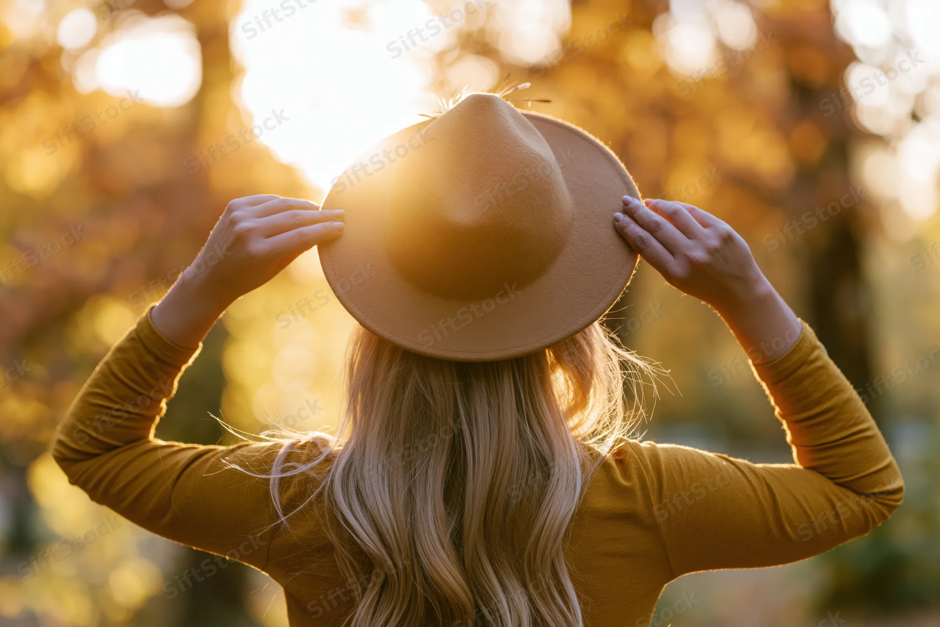 Woman from behind holding a felt hat in autumn sunlight among trees