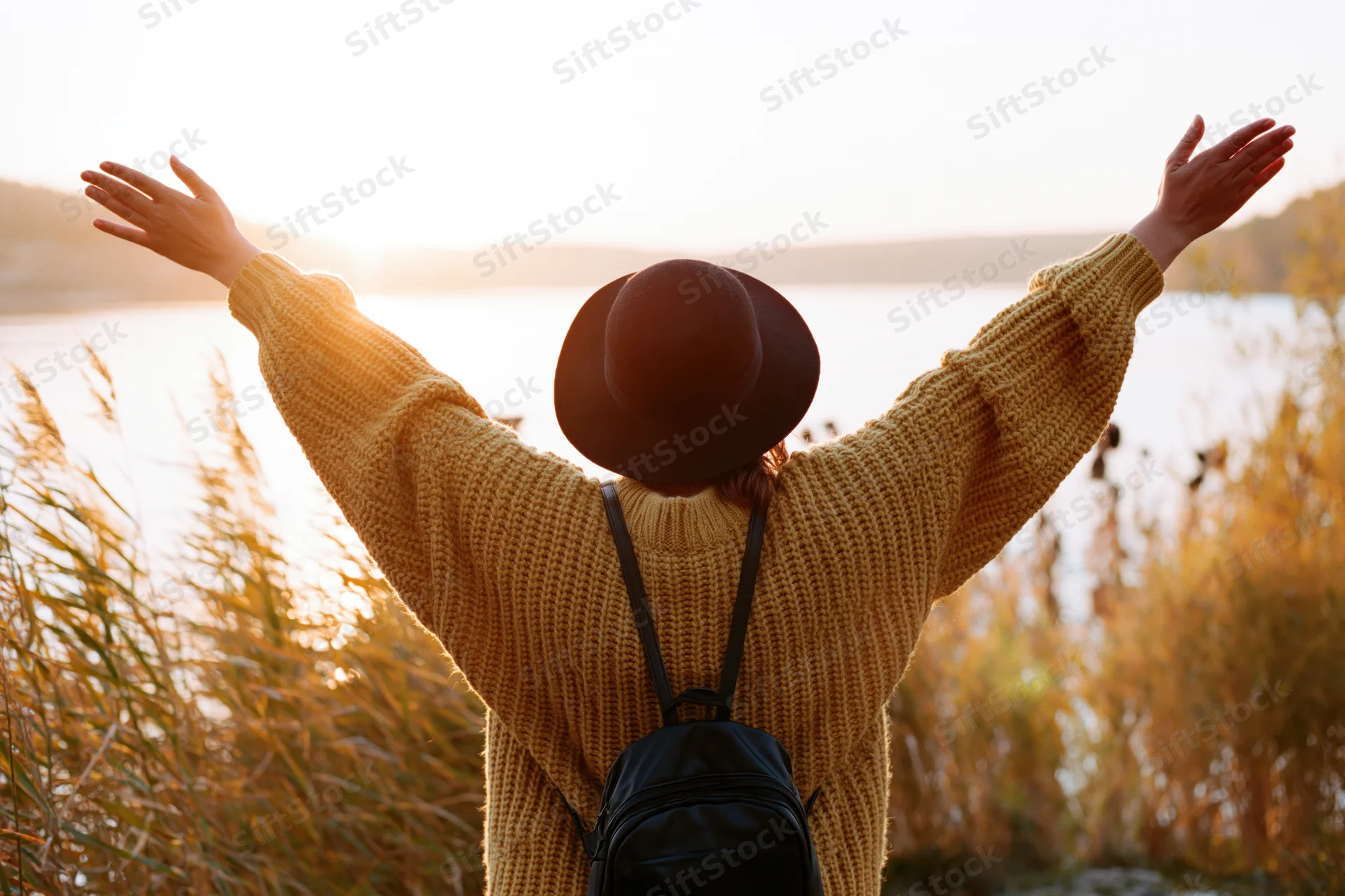Woman with arms raised facing sunset over lake, wearing hat and sweater, backpack on back, surrounded by reeds